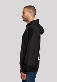 Černá mikina s kapucí a přední kapsou, oversized střih, texturovaná tkanina a barevný grafický design na zádech. Nosit s modrými džíny.