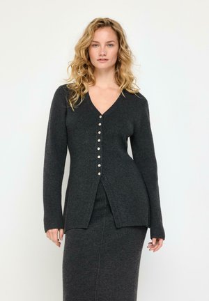 Donna bionda che indossa un cardigan grigio scuro con bottoni e gonna lunga abbinata, in piedi davanti a uno sfondo bianco semplice.