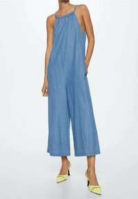 Combinaison en denim bleu avec jambes larges, décolleté halter et détail froncé. Associée à des chaussures à talons hauts jaunes.