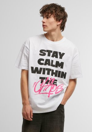 Hvid bomulds t-shirt med fed sort tekst "STAY CALM WITHIN THE CHAOS" og en pink spraymaler-lignende accent. Løs pasform med korte ærmer.