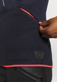 Marine blauwe fleece kledingstuk met een ritszak. Voorzien van een felrode voering en contrasterende rode bies. Inclusief een zwart logo-embleem.