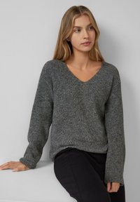 Jonge vrouw met lang blond haar, gekleed in een grijze gebreide V-halstrui en zwarte broeken, zittend met één hand op een witte oppervlakte.
