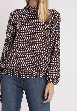 Langärmliges Top mit hohem Kragen in Bordeauxrot, mit einem weißen diagonalen Rautenmuster. Verfügt über einen elastischen Saum und lockere Passform, kombiniert mit blauen Jeans.