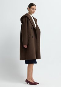 Brauner Wollmischungsmantel mit Kapuze, doppelt geknöpftem Design, großen Vordertaschen und schwarzen Knöpfen. Kombiniert mit einem dunkelblauen Jeansrock und burgunderfarbenen Schuhen.