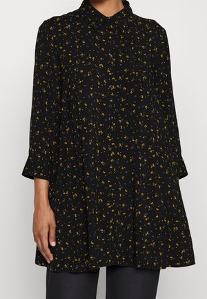 Zwarte tuniek met lange mouwen en knopen, bedrukt met een kleine verspreide gele en grijze bloemenprint, gedragen over zwarte broek.