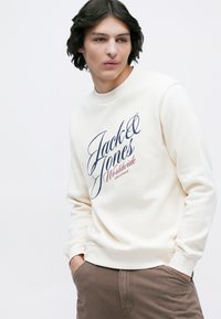 Mladý muž s tmavými vlasmi, oblečený v krémovej mikine s nápisom "Jack & Jones Worldwide Originals", hnedých nohaviciach, s rukami v vreckách a pozerajúci sa doľava.