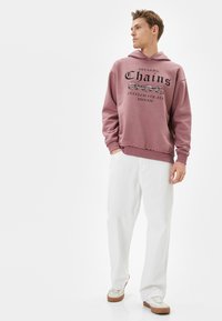 Maroon hoodie med grafiken "Breaking Chains" och vit text, parat med lösa vita byxor och ljusa sneakers. Slät tygtextur.