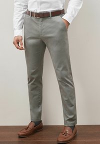 Pantalons gris clair avec une finition texturée, jambe fuselée, poches latérales et une ceinture en cuir marron. Associé à des mocassins marron.