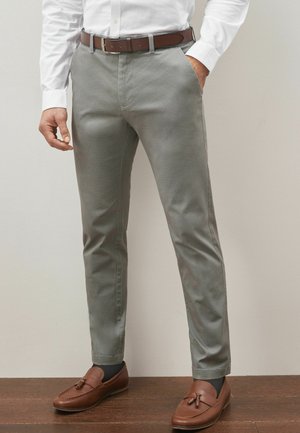 Pantalones gris claro con un acabado texturizado, pierna ajustada, bolsillos laterales y un cinturón de cuero marrón. Combinados con mocasines marrones.