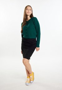 Cardigan corto in maglia a trecce verde, gonna nera aderente e sneaker alte gialle con lacci bianchi, in piedi contro uno sfondo neutro.