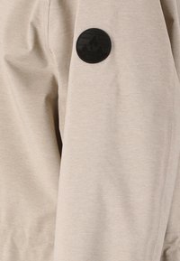 Veste beige avec une texture lisse, arborant un patch logo circulaire noir sur la manche gauche. Design simple sans motifs supplémentaires.