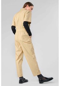 Combinaison beige à manches courtes avec une taille élastique, deux poches sur les côtés et un col. Associée à des chaussettes noires texturées et des mocassins brillants.