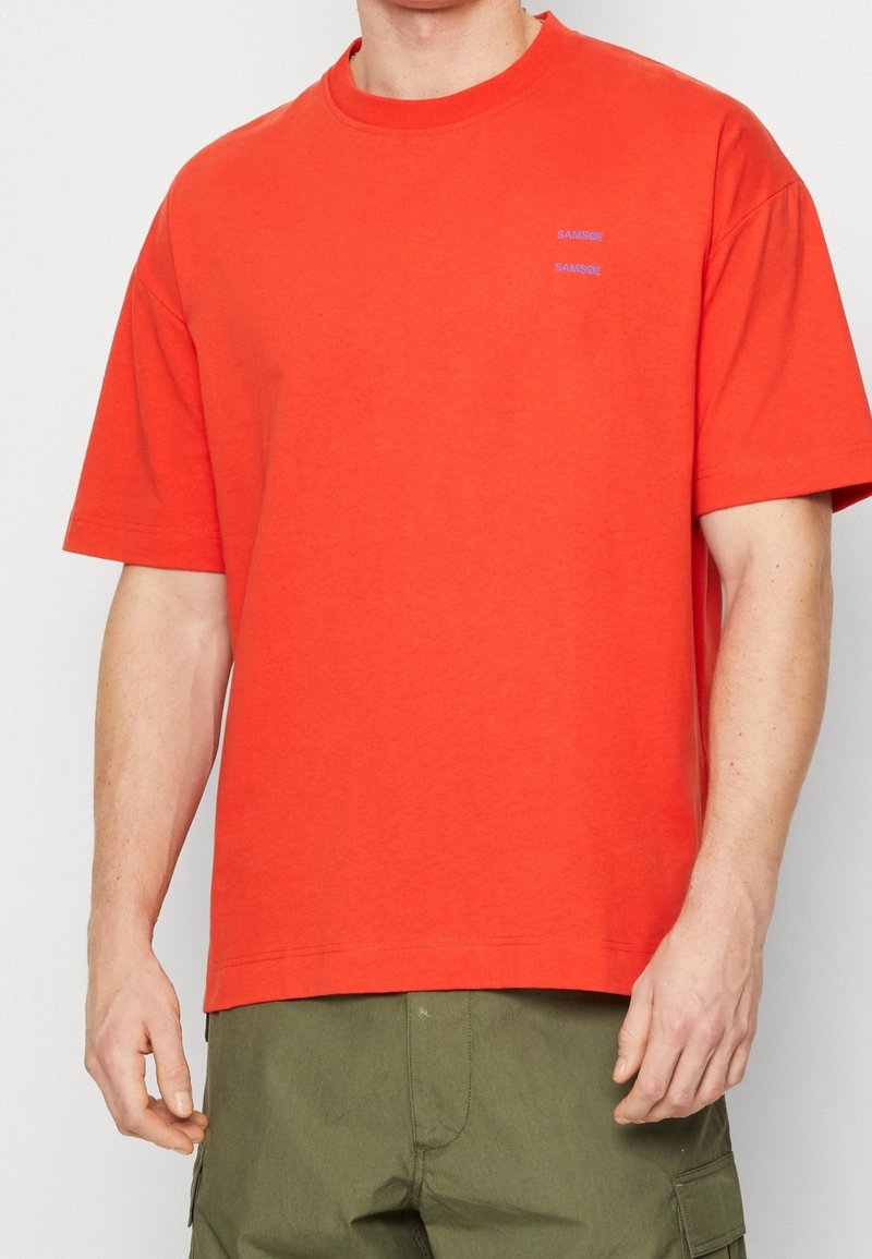 T-shirt en coton rouge vif avec une coupe décontractée, des manches courtes et un col rond. Présente un petit logo "SAMSØE" violet sur la poitrine.