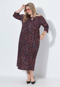 Nainen, jolla on päällään tumma paisleymuotoinen pitkä mekko, mustat korkokengät, oranssit aurinkolasit, kultaketju ja korvakorut, poseeraa valkoista seinää vasten.