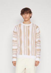 Beige och vit mönstrad stickad tröja med rund halsringning, med vertikala ränder och texturerade mönster, som bärs med ljusa byxor.