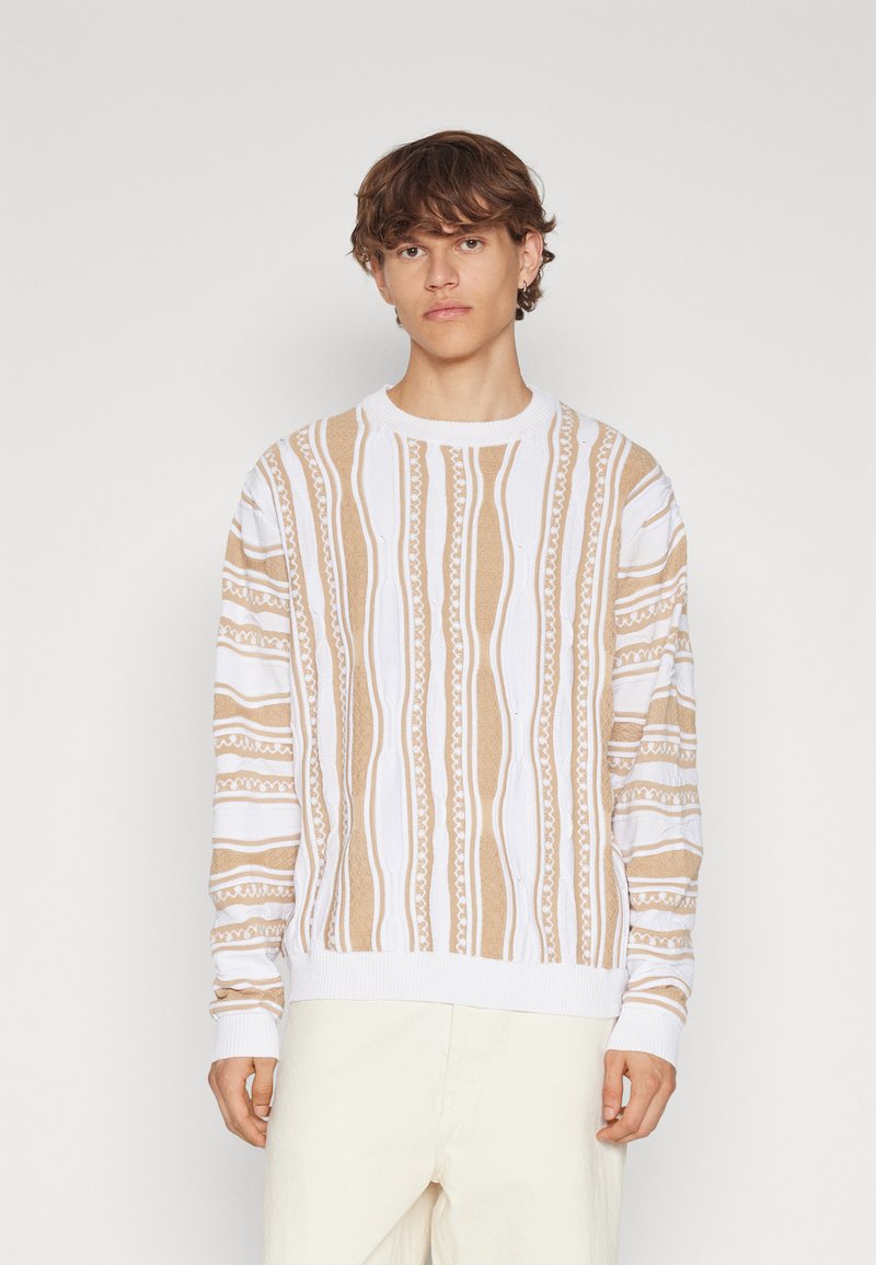 Beige och vit mönstrad stickad tröja med rund halsringning, med vertikala ränder och texturerade mönster, som bärs med ljusa byxor.