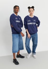 Námornícke tričká s dlhým rukávom, s nápisom "KONKURENCIA" v bielej farbe. Model naľavo nosí široké denimm šortky; napravo je v priliehavých džínsach. Čierno-biele tenisky.
