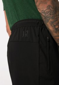 Pantalones deportivos negros con una textura suave, cinturilla elástica, bolsillo lateral con cremallera y un logo discreto en la parte posterior. El diseño es funcional y estilizado.