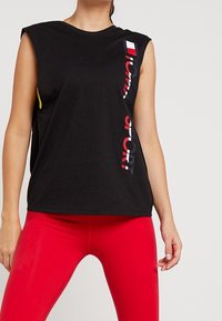Donna che indossa un top senza maniche nero con il logo verticale "Tommy Sport" e leggins rossi vivaci, in piedi con una mano rilassata al suo fianco.