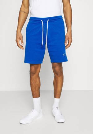 Mann trägt leuchtend blaue Shorts mit Kordelzug, weiße Socken, weiße Sneakers und ein weißes ärmelloses Oberteil vor schlichtem Hintergrund.