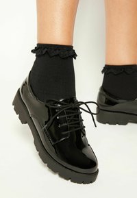 Scarpe in pelle verniciata nera con punta arrotondata, design con lacci e suola spessa in gomma. Indossate con calzini neri a balze.