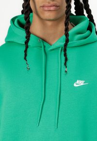 Zaļš kapucis, izgatavots no mīksta auduma, ar kangurū kabatu, auklām un baltu Nike logotipu uz krūtīm.