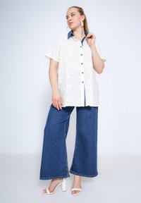 Witte floral kant blouse met een blauwe denim kraag, knoopsluiting, en wijde donkerblauwe jeans, gecombineerd met witte haksandalen.
