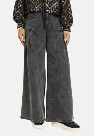 Persona que lleva pantalones vaqueros negros desteñidos de pierna ancha con detalles visibles de costuras y zapatillas negras con suelas blancas.