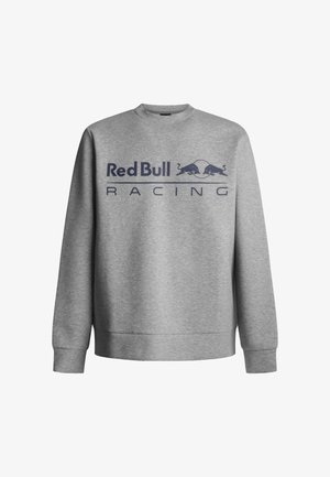 Mann trägt ein schlichtes graues Langarm-Sweatshirt und blaue Jeans, steht mit dem Rücken zur Kamera vor grauem Hintergrund.