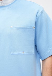 T-shirt de mangas curtas de cor azul claro, fabricado em tecido macio, com um grande bolso no peito com botões de pressão e decote redondo.