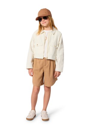 Jongere die een beige pet en korte broek draagt, een crèmekleurig jack, wit shirt, zonnebril en witte sneakers, staand in een ontspannen houding.