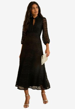 Schwarzes Spitzen-Midikleid mit hohem Kragen, durchsichtigen langen Ärmeln und schwingendem Rock. Das Model hält eine hellfarbige Clutch. Silberne Knöchelriemen.