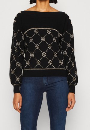 Femme portant un pull en maille noir avec un motif géométrique doré et logo, détails de boutons sur les épaules, associé à un jean bleu.