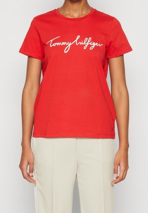 Lyhythihainen punainen T-paita, jossa on valkoinen "Tommy Hilfiger" -logo, yhdistettynä vaaleanbeigeihin housuihin seisovalla henkilöllä.