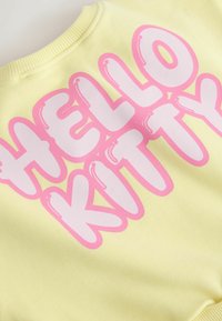 Tela amarilla con texto burbuja grande en rosa y blanco que deletrea "HELLO KITTY" en la parte delantera.