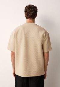 Beige textured short-sleeve shirt with a loose fit, featuring a square pattern throughout and a round neckline.