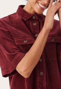 Bordeauxrotes Cordhemd mit einem Kragen, großen Knöpfen und kurzen Ärmeln. Verfügt über vertikale Rippenstrukturen und eine Brusttasche als Detail.
