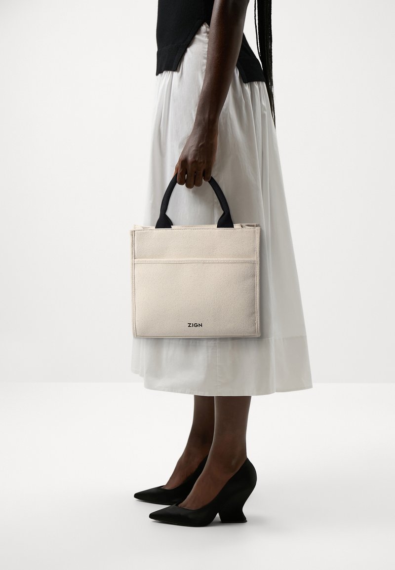 Femme en jupe blanche et top noir tenant un sac à main rectangulaire beige avec des poignées noires et des talons hauts noirs, debout sur un fond blanc.