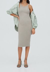 Robe midi grise sans manches à coupe ajustée, associée à une chemise oversize boutonnée vert clair et des sandales à talons vert olive.