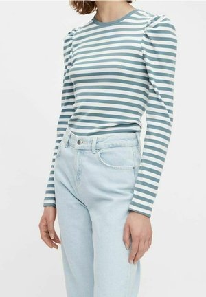 Nainen pukeutunut pitkähihainen, vartalonmyötäinen paita, jossa on turkoosin ja valkoisen vaakaraidat, yhdistettynä vaaleansinisiin korkeavyötäröisiin farkkuihin.