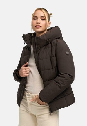 Frau trägt eine dunkelbraune wattierte Jacke mit Kapuze über einem hellbeigen Pullover und cremefarbenen Hosen, steht vor einem einfarbigen Hintergrund.