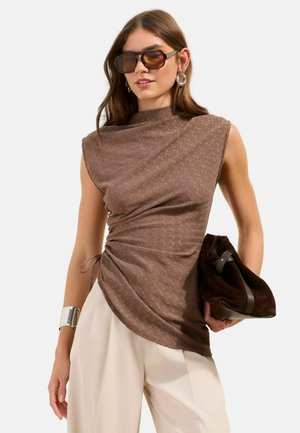 Žena oblečená v hnedomá textúrovanom tielku bez rukávov s bočnými záhybmi, krémových širokých nohaviciach, veľkých slnečných okuliaroch, striebornom náramku a držiaca tmavohnedú kabelku.