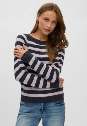 Mujer posando con un jersey de rayas negras y gris claro y unos vaqueros azules sobre un fondo blanco liso.