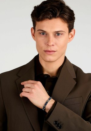Blazer marrone con rever a punta, indossato sopra una camicia nera. Un braccialetto argento e blu con link rettangolari è visibile sul polso destro.