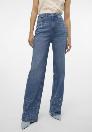 Blå højtaljede viddejeans med spredte små hvide studser på øverste hofter og nederste ben, båret med lyseblå spidse sko.