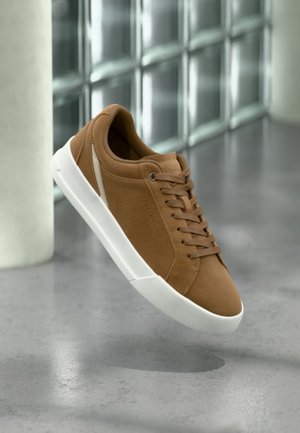 Bruine suède sneaker met witte zool en veters, zwevend boven een gepolijste betonnen vloer in een moderne binnenomgeving.