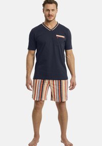 Marineblaues Baumwoll-V-Ausschnitt-T-Shirt mit einer gestreiften Tasche; kombiniert mit mehrfarbigen gestreiften Shorts in Orange, Marineblau und Weiß. Barfußhaltung.
