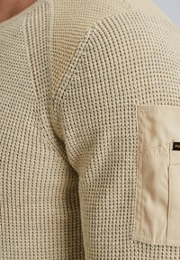 Beżowy sweter robiony na drutach z teksturowanym wzorem, z naszywaną kieszonką na lewym ramieniu, wykonany z miękkiego, przewiewnego materiału.