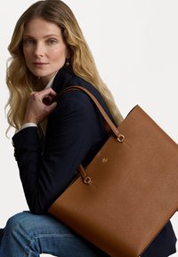 Bolsa tote de couro castanho com um design estruturado, apresentando detalhes em dourado e alças duplas. Superfície texturizada com um destaque de logotipo.