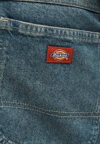 Tissu en denim bleu avec un patch rectangulaire rouge du logo Dickies cousu près des coutures des poches.